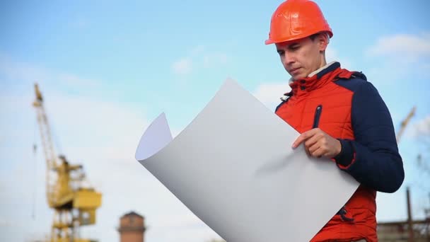 Чоловік архітектор портрет будівельника в помаранчевому шоломі дивиться інструменти проект проти блакитного неба і будівлі — стокове відео