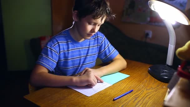 Teenage boy writes in notebook homework sitting at lamp table  evening room — Stock Video