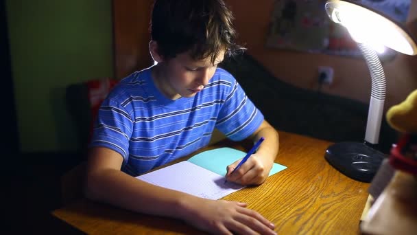 十代の少年は、テーブルランプの夜の部屋に座ってノートの宿題で書きます — ストック動画
