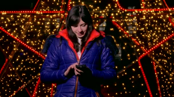 Ragazza donna in possesso di un telefono intelligente guardando i social media la notte dietro bokeh luce invernale — Video Stock