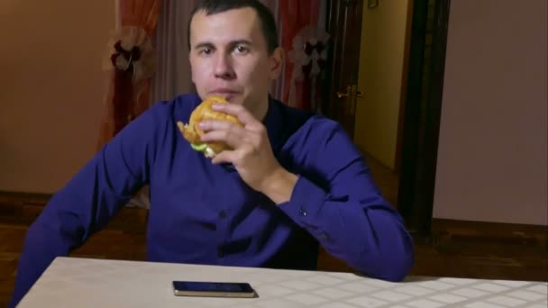 Hombre comiendo una hamburguesa con un teléfono inteligente sentado en un restaurante de comida rápida — Vídeo de stock