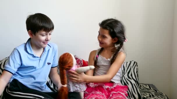 Niña y niño jugando con una muñeca sentada en una cama — Vídeo de stock