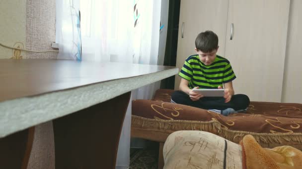 Мальчик-подросток играет на планшете, сидя на диване — стоковое видео