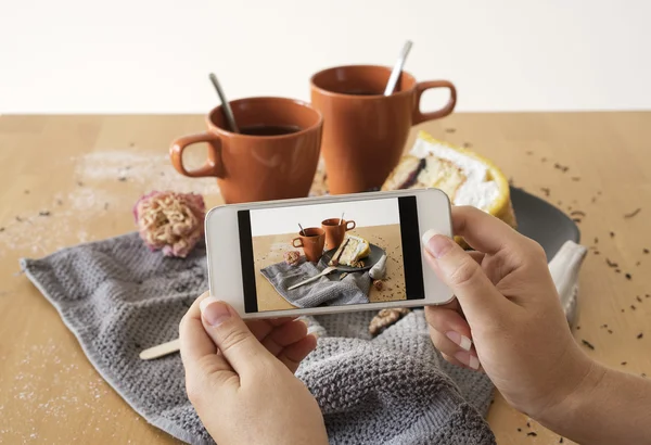 She makes food photo on his phone. The composition of the two cups of tea with cake , nuts , flowers and cloth.