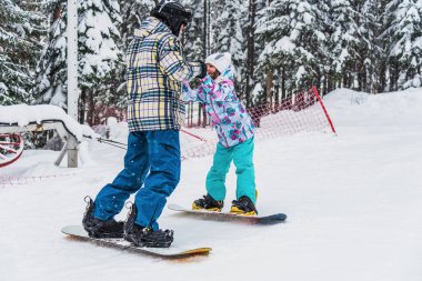 Babası kızına snowboard yapmayı öğretiyor. Birlikte dağdan aşağı iniyorlar.