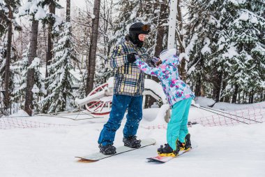 Babası kızına snowboard yapmayı öğretiyor. Birlikte dağdan aşağı iniyorlar.