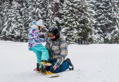 Baba, kızına bir kış günü kar kayağı bağlayıcısını kayak yamacına bağlamasında yardım eder.