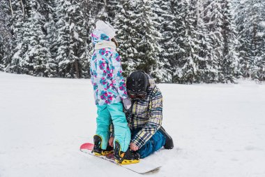 Baba, kızına bir kış günü kar kayağı bağlayıcısını kayak yamacına bağlamasında yardım eder.