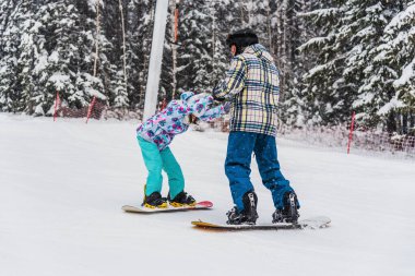 Baba kızına snowboard yapmayı öğretiyor. Kayak yamacından aşağı yuvarlanırlarken ellerini tutuyor.