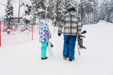Baba kızına snowboard yapmayı öğretiyor. Birlikte kayak pistine çıkıyorlar.