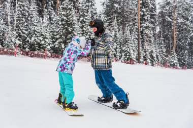 Baba kızına snowboard yapmayı öğretiyor. Kayak yamacından aşağı yuvarlanırlarken ellerini tutuyor.