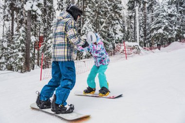 Baba kızına snowboard yapmayı öğretiyor. Kayak yamacından aşağı yuvarlanırlarken ellerini tutuyor.