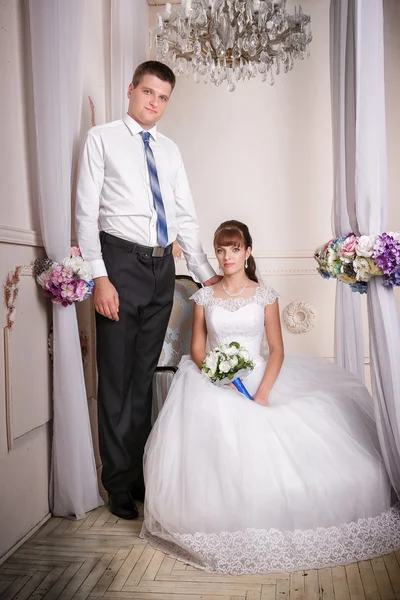 The bride sits in a chair and the groom stands near bride in the room with a beautiful interior — Stock Photo, Image