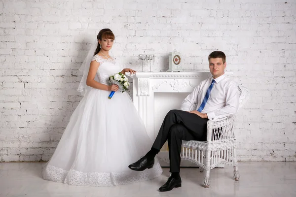 El novio está sentado en una silla y la novia está de pie cerca de la chimenea en el fondo de la pared de ladrillo blanco —  Fotos de Stock