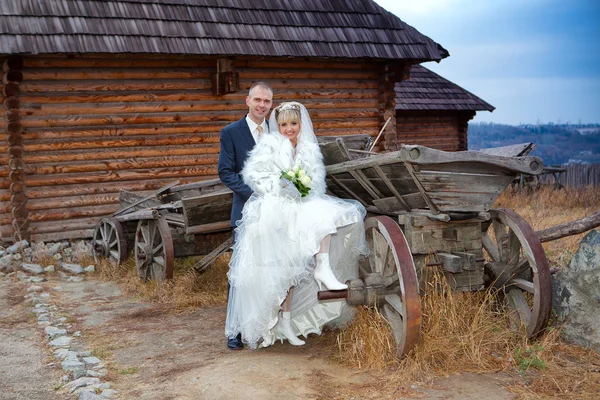 O noivo e a noiva contra a casa de madeira e um velho carrinho de cavalo — Fotografia de Stock