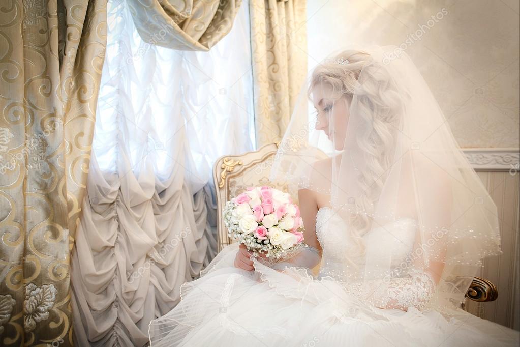 Portrait of the bride with a wedding bouquet from roses in the hand sitting on a sofa near a window