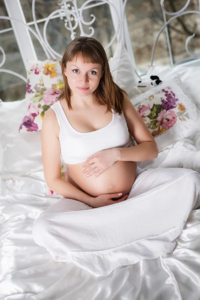 Beautiful pregnant woman with children's bootees posing while siting on a bed at home — Stock Photo, Image