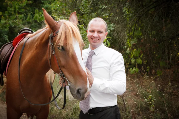 Novio durante el paseo en el día de su boda contra un caballo marrón — Foto de Stock