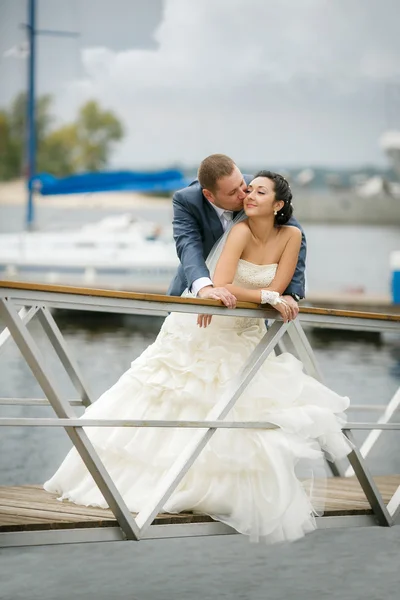 Pareja joven enamorada de la novia y el novio con un ramo posando en un muelle de fondo con yates, el día de la boda en el verano . — Foto de Stock