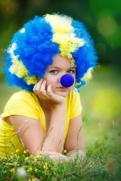 Meisje in clown pruik met blauwe neus ligt op het groene gras in het park — Stockfoto