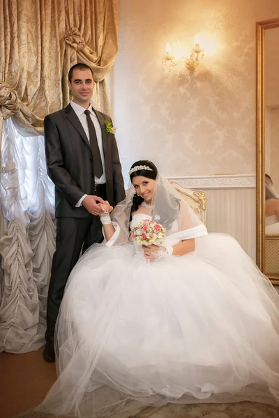 The bride sits in a chair and the  groom stands near groom in the room with a beautiful interior — Stock Photo, Image