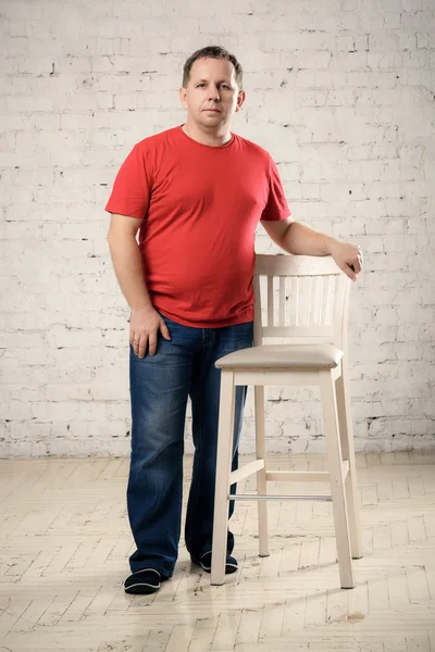 Homem em uma camiseta vermelha fica perto de uma cadeira em estúdio — Fotografia de Stock