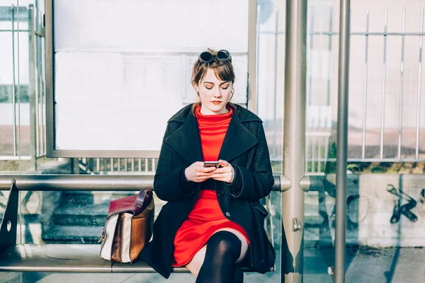 Vrouw zitten bij bushalte via smartphone — Stockfoto