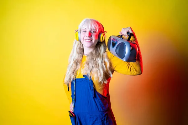 Young beautiful caucasian woman dancing indoor on yellow background holding vintage stereo - Isolated diverse female clubbing holding vintage radio - happiness, excitement, dancing concept