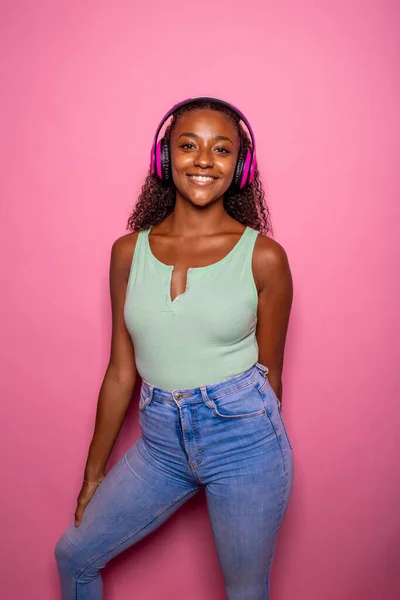 ピンクの背景で陽気な黒人女性が音楽を聞いているスマートフォンを保持 ヘッドフォンを身に着けている電話を使用して独立した黒の若い女の子 ストリーミング 音楽コンセプト — ストック写真