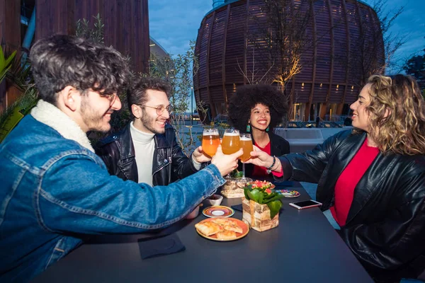 Group of four multiethnic friends sitting outdoor club drinking beer making toast celebrating happy positive moments together having fun