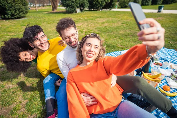 四个多种族朋友组成的小组在户外用智能手机自娱自乐 一起在公园里笑着野餐 — 图库照片