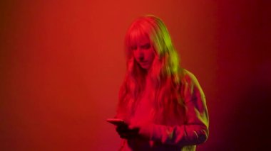 Kırmızı arka planda, kameraya bakan akıllı telefon kullanan, sarışın, beyaz bir kadının stüdyo fotoğrafı.