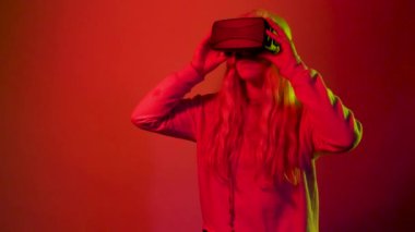 Kırmızı arka planda beyaz bir kadının VR gözlük taktığı ve arttırılmış gerçeklikte fütürist kulaklık taktığı bir fotoğraf.