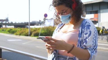 Ağır çekimde, çok ırklı, maskeli, bluetooth kulaklıklı ve akıllı telefon kullanan genç bir kadın.