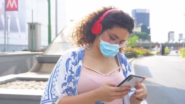 Dışarıda maskeli, bluetooth kulaklıklı ve akıllı telefon kullanan genç bir kadın.