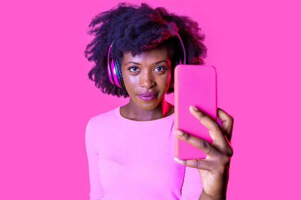 若い美しい黒人女性がピンクの背景を見ながら自撮り 孤立したアフリカの若い女性がヘッドフォンスタジオショットを着てライブストリーミング ソーシャルネットワーク 多様性の概念 — ストック写真