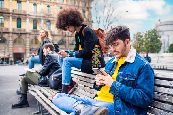 Четыре мультиэтнических человека незнакомцы или друзья сидят на скамейке запасных с помощью смартфона игнорируя зависимость от социальных тенденций после использования смайликов