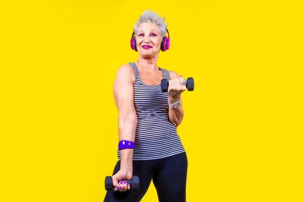 Elderly woman training using dumbbells on yellow background stretching using gym weight listening music using bluetooth wireless headphones