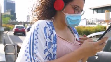 Çok ırklı genç bir kadın dışarıda koruyucu maske takıyor. Cep telefonu ve kablosuz kulaklık kullanıyor. Haberleri arıyor ve internette sohbet ediyor.