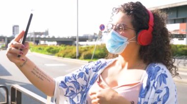 Ağır çekimde, açık havada cerrahi maske takan genç bir ırk kadını akıllı telefon ve kablosuz bluetooth kulaklıkla video görüşmesi yapıyor.