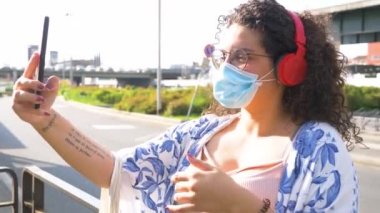 Açık havada cerrahi maske takan genç bir ırk kadını elinde akıllı telefon ve kablosuz bluetooth kulaklıkla video görüşmesi yapıyor.