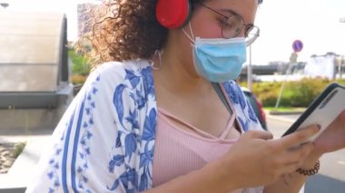 Yavaş çekim genç kadın dışarıda koruyucu maskeli sörf ağı takıyor cep telefonu ve kablosuz kulaklık kullanıyor haberleri arıyor ve internette sohbet ediyor.