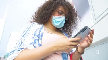 Çok ırklı genç bir kadın koruyucu maskeli sörf ağı takıyor şehirde cep telefonu kullanıyor, haberleri arıyor ve arkadaşlarıyla sohbet ediyor.