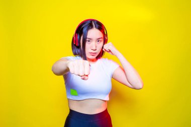 Asyalı genç bayan boks antrenmanı yapıyor sarı arka planda tutum ve özgüvenle müzik dinliyor.