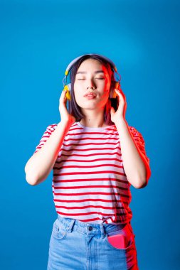 Sakin Asyalı kadın mavi arka planda izole edilmiş bluetooth kulaklıklı ve gözleri kapalı müzik dinliyor. 