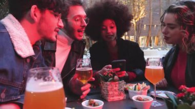 Dört etnik gruptan oluşan bir grup dışarıda oturup akıllı telefon kullanarak birlikte mutlu anları kutluyor ve birlikte eğleniyorlar.