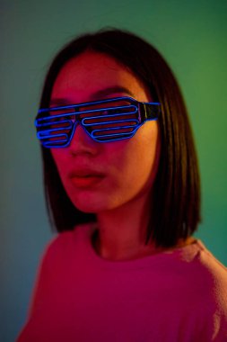 Kulaklık takan ve yeşil arka planda fütürist gözlükler takan genç bir kadın, izole edilmiş bir kadın, sessiz diskoda gidiyor, neon gözlük takıyor, kulaklık takıyor, parti, gelecek, teknoloji konsepti. 