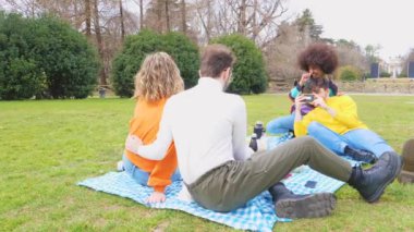 Farklı etnik çeşitlilikte dört neşeli arkadaş parkın dışında piknik yaparken akıllı telefon kullanarak fotoğraf çekerken hatıralar yaratırken eğleniyorlar.