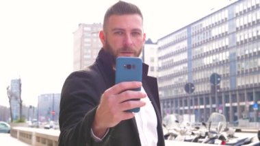 Yavaş çekim genç sakallı beyaz iş adamı gibi dışarıda akıllı telefon kullanan kameraya pozitif ve kendinden emin bakarak selfie çeken. 