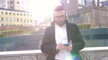 Ağır çekimde, beyaz sakallı, açık havada müzik dinleyen kulaklıklı, akıllı telefon saplı, gülümseyen ve gevşemiş genç bir adam.